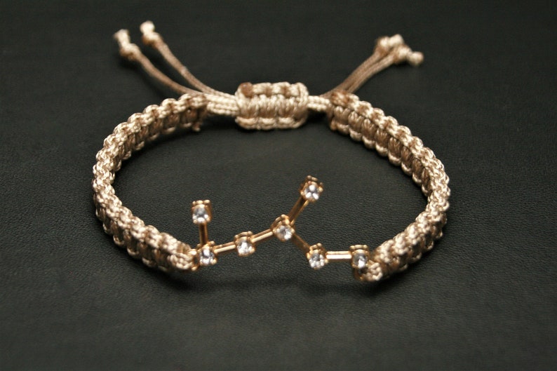 Peut inclure: Un bracelet tress&eacute; beige avec un charme de constellation Vierge en or avec des strass clairs.