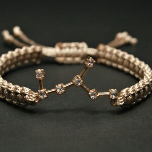 Peut inclure: Un bracelet tress&eacute; beige avec un charme en or repr&eacute;sentant une constellation de petits strass transparents.