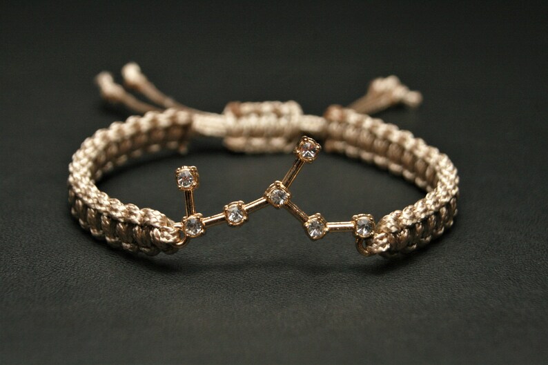 Peut inclure: Un bracelet tress&eacute; beige avec un charme en or repr&eacute;sentant une constellation de petits strass transparents.