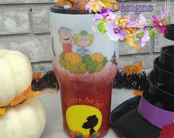 May include: A red and white glitter tumbler with a Peanuts Halloween design. The tumbler features Charlie Brown, Lucy, and Linus with pumpkins. The text "There he is!" is printed on the bottom of the tumbler.