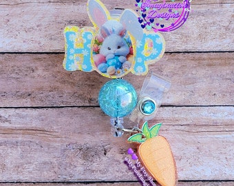 May include: A glittery Easter-themed badge reel with a bunny and carrot charm. The word "HOP" is spelled out in blue letters with a floral pattern. The bunny is white and blue, and the carrot is orange.