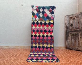May include: A long, narrow rug with a diamond pattern in shades of pink, red, yellow, blue, and black. The rug is made of wool and has a hand-woven texture.