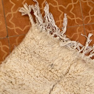 May include: A white fluffy rug with fringe on the edges. The rug is lying on a brown tiled floor with a geometric pattern.