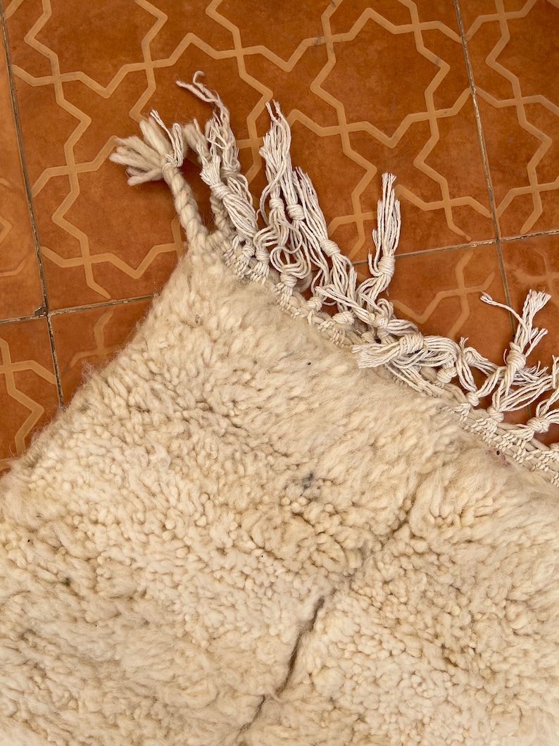 May include: A white fluffy rug with fringe on the edges. The rug is lying on a brown tiled floor with a geometric pattern.