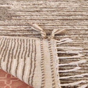 May include: A beige and brown textured rug with a geometric pattern. The rug has a fringe on one edge and a folded corner revealing the underside with vertical stripes. The rug is made of a soft, plush material.