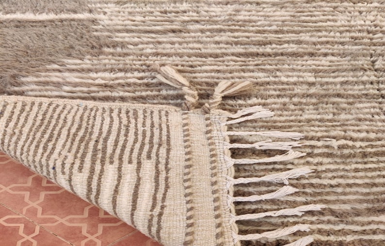 May include: A beige and brown textured rug with a geometric pattern. The rug has a fringe on one edge and a folded corner revealing the underside with vertical stripes. The rug is made of a soft, plush material.