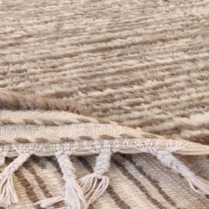 May include: Close-up of a beige and brown textured rug with a striped pattern. The rug has a fringe of knotted tassels along one edge. The fibers appear soft and plush, creating a cozy aesthetic.