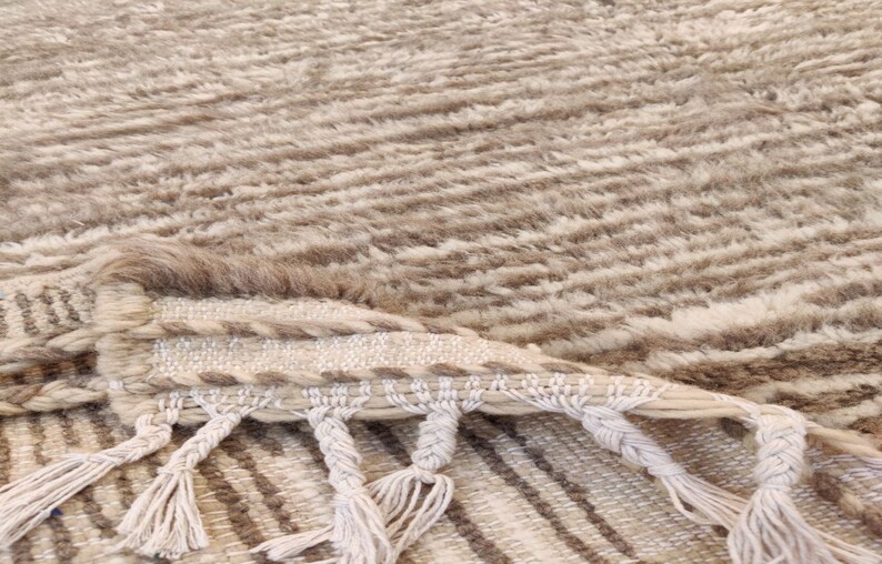 May include: Close-up of a beige and brown textured rug with a striped pattern. The rug has a fringe of knotted tassels along one edge. The fibers appear soft and plush, creating a cozy aesthetic.