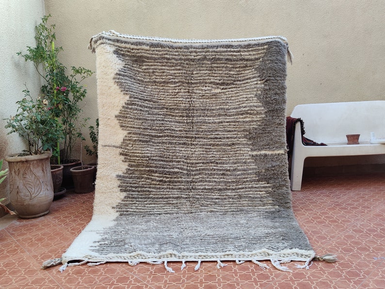 May include: A rectangular, hand-woven rug with a gradient design. The rug transitions from white to gray and brown, with horizontal stripes. Tassels adorn the bottom edge. This rug is suitable for home decor.