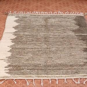 May include: Rectangular rug with a textured, striped design in shades of gray and off-white. The rug has a fringe along the edges and is suitable for home decor. The rug is placed on a terracotta tile floor.