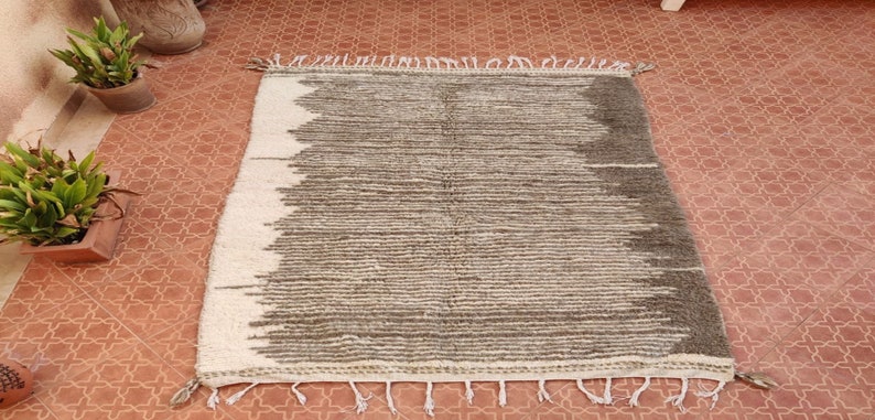 May include: Rectangular rug with a textured, striped design in shades of gray and off-white. The rug has a fringe along the edges and is suitable for home decor. The rug is placed on a terracotta tile floor.