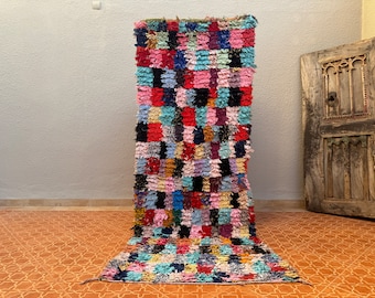 May include: A colourful Moroccan rug with a chequered pattern in shades of pink, blue, red, and black. The rug is made of woven wool and has a fringed edge.