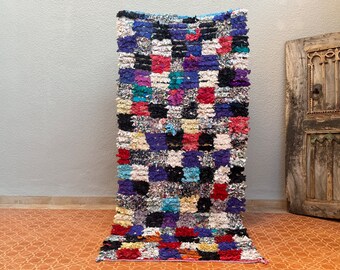 May include: A colourful, handwoven Moroccan rug with a chequered pattern in shades of blue, red, white, black, and purple. The rug is made of recycled materials and has a unique, textured look.