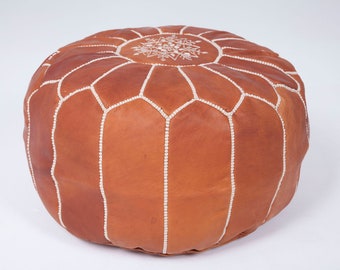 May include: Round, brown leather pouf with white stitching. The top features a white geometric design. This decorative ottoman is a stylish accent piece for any living space, offering both comfort and visual appeal.