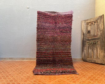 May include: A long, rectangular rug with a woven texture. The rug has a mix of pink, red, brown, and green colours. The rug is lying on a tiled floor.