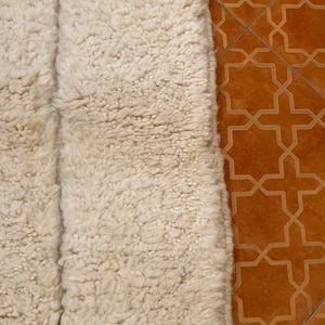 May include: Close-up of a white fluffy rug with a textured surface. The rug is placed on a brown tiled floor with a geometric pattern.