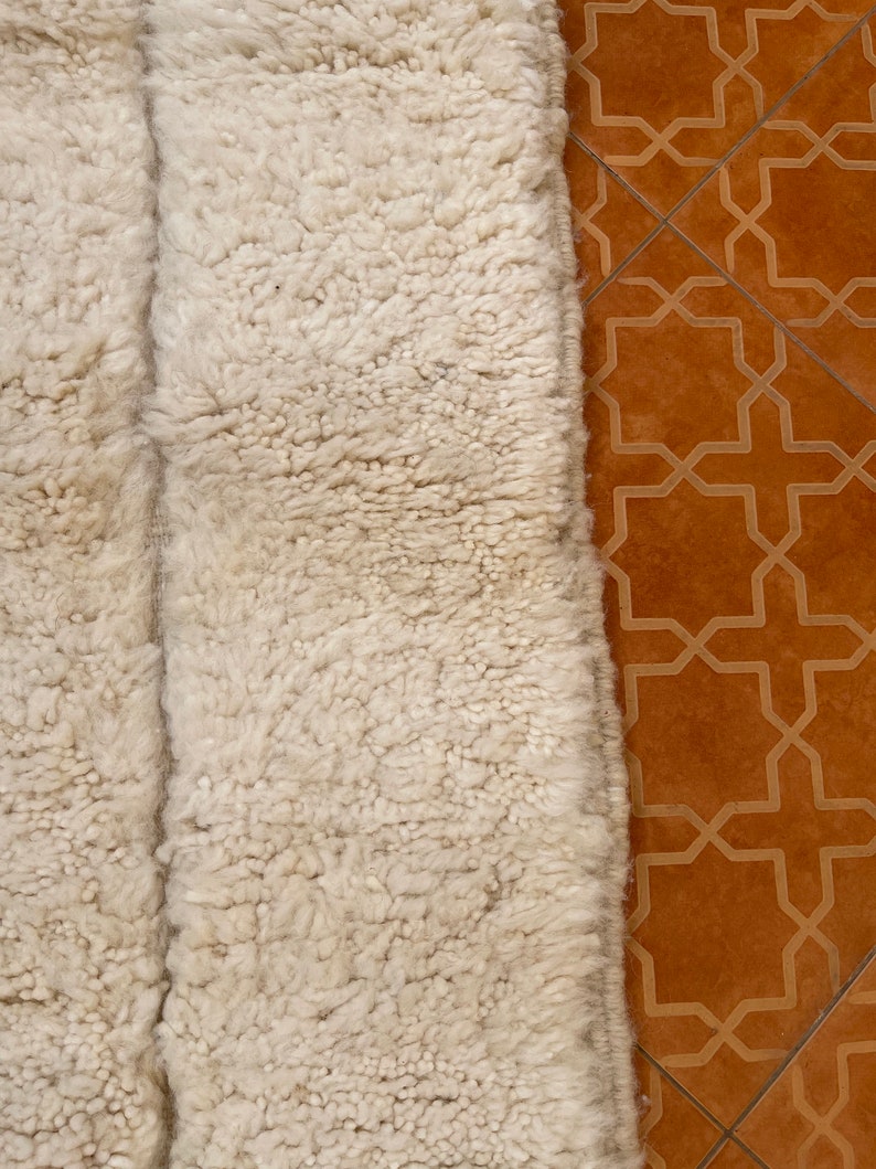 May include: Close-up of a white fluffy rug with a textured surface. The rug is placed on a brown tiled floor with a geometric pattern.