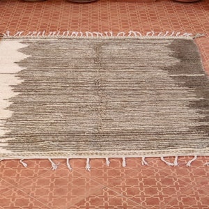 May include: A rectangular, hand-woven rug with a striped design. The rug features a gradient pattern, transitioning from white to shades of gray and brown. The edges are finished with tassels.