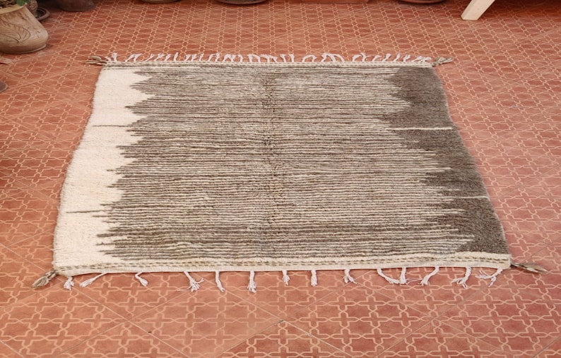 May include: A rectangular, hand-woven rug with a striped design. The rug features a gradient pattern, transitioning from white to shades of gray and brown. The edges are finished with tassels.