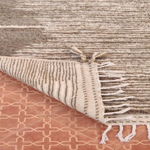 May include: A beige and gray textured rug with a geometric design. The rug has a soft, plush appearance with a striped pattern and fringe detailing on the edge. The rug is partially folded, revealing a contrasting underside.