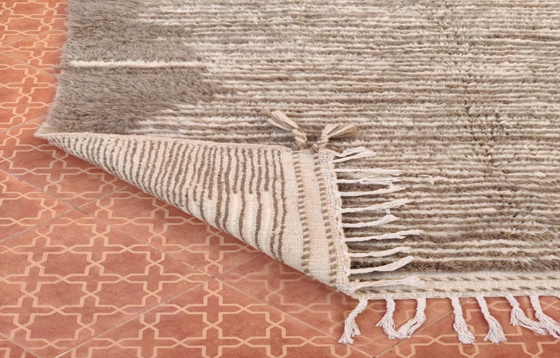May include: A beige and gray textured rug with a geometric design. The rug has a soft, plush appearance with a striped pattern and fringe detailing on the edge. The rug is partially folded, revealing a contrasting underside.