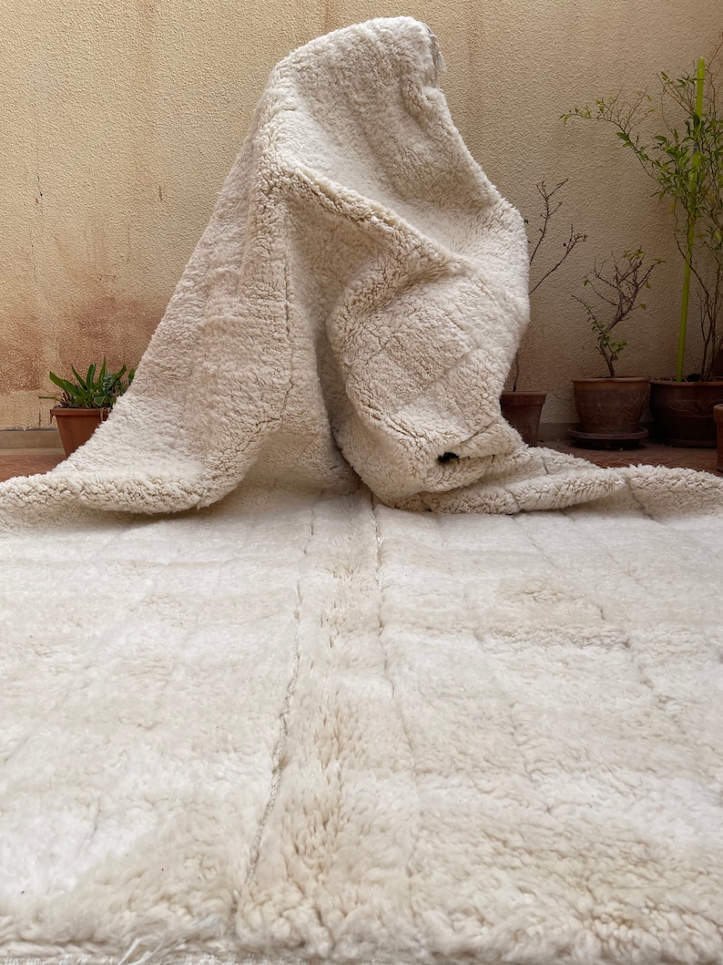 May include: A white sheepskin rug with a fluffy texture. The rug is folded in half and is lying on a tiled floor.