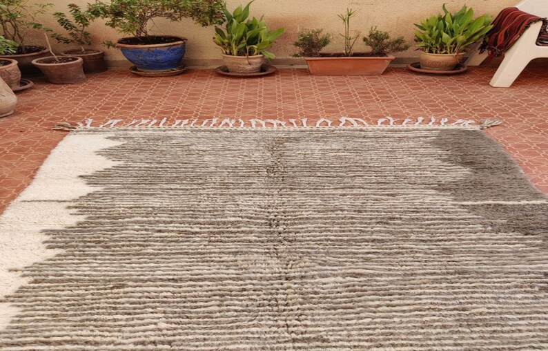 May include: A rectangular, hand-woven rug with a textured, striped pattern. The rug features a color scheme of white, gray, and brown. The rug has a fringe on one end and is suitable for indoor use.