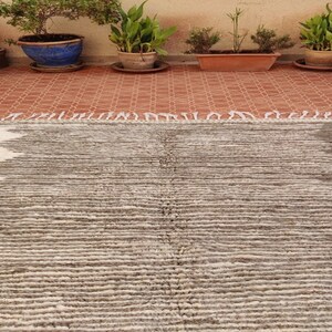 May include: A rectangular, hand-woven rug with a textured, striped pattern. The rug features a color scheme of white, gray, and brown. The rug has a fringe on one end and is suitable for indoor use.