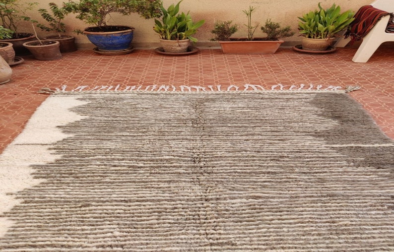 May include: A rectangular, hand-woven rug with a textured, striped pattern. The rug features a color scheme of white, gray, and brown. The rug has a fringe on one end and is suitable for indoor use.