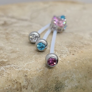 May include: A white body jewelry barbell with three clear and colorful gem accents. The barbell has a pink gem at the bottom, a blue gem in the middle, and a clear gem at the top. The barbell is on a light brown rock.