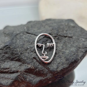 May include: Silver body jewelry charm featuring a minimalist line drawing of a face.
