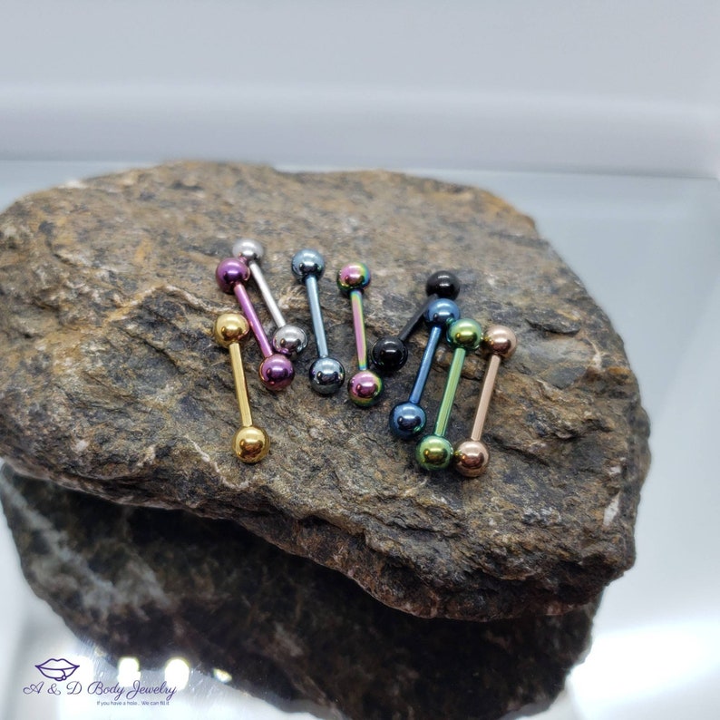 May include: A collection of 10 colorful barbell body jewelry pieces with different colored balls on a rock surface. The barbells are made of surgical steel and have a variety of colors, including gold, silver, black, blue, green, and pink.