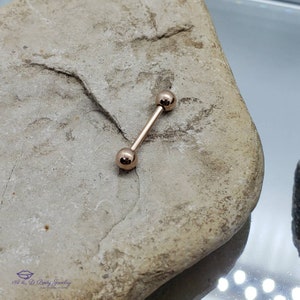 May include: A rose gold barbell with two round balls on each end. The barbell is lying on a light brown rock.