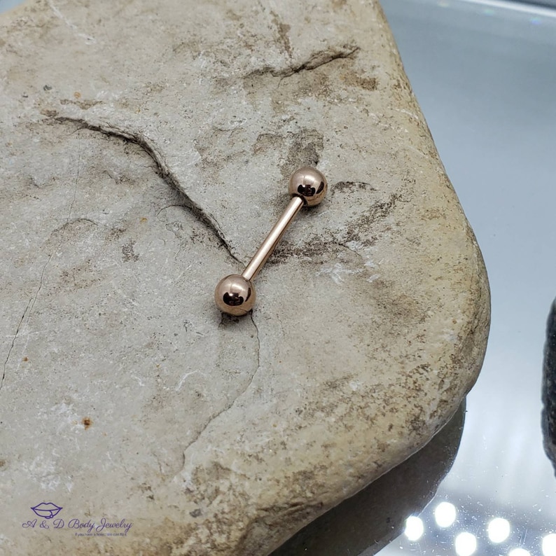 May include: A rose gold barbell with two round balls on each end. The barbell is lying on a light brown rock.