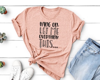 May include: A peach-coloured t-shirt with the text "Hang on. Let me overthink this..." in black. The shirt is styled with light blue jeans and white trainers. Small potted succulents are arranged around the clothing.