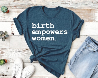 May include: A teal blue t-shirt with the text "birth empowers women." The shirt is laid flat on a white wood surface with a pair of white trainers, a pair of light blue jeans, and three small potted succulents.