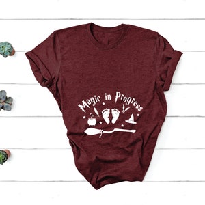 May include: Burgundy t-shirt with a white graphic that says "Magic in Progress" with a broom, cauldron, and baby footprints.