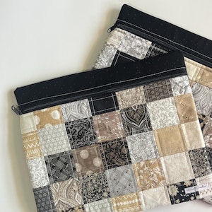 May include: Two quilted fabric pouches with a patchwork design. The pouches feature a black zipper and a black top border. The patchwork squares are in shades of beige, gray, and black, with various patterns.