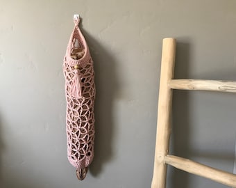 May include: A pink crocheted hanging storage bag with a wooden bead and tassel. The bag is hanging on a white hook on a light gray wall. A wooden ladder is in the background.