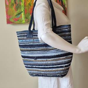 Może przedstawiać: Dżinsowa torba typu tote z poziomymi paskami w odcieniach niebieskiego, białego i brązowego. Torba ma ciemnoniebieskie uchwyty i jest prezentowana na manekinie. Torba ma około 38 cm szerokości i 30 cm wysokości.