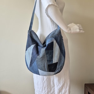 Puede incluir: Un bolso de hombro de patchwork de mezclilla con una correa larga. El bolso está hecho de varios tonos de mezclilla azul, creando un patrón geométrico. El bolso se exhibe en un maniquí con un vestido blanco.