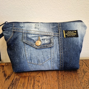 Op de afbeelding: Een denim-achtige pouch met een ritssluiting en een klein vakje. De pouch is gemaakt van blauwe denim stof en heeft een gouden knoop en een zwart label met de tekst "Authentic Crafted for Comfort".