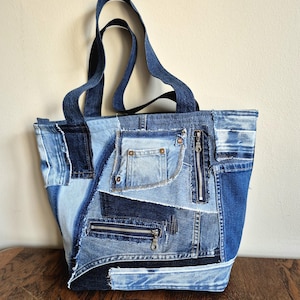 May include: A tote bag constructed from various shades of blue denim fabric patches. The bag features two shoulder straps, a visible pocket with copper-colored rivets, and two functional metal zippers integrated into the patchwork design.