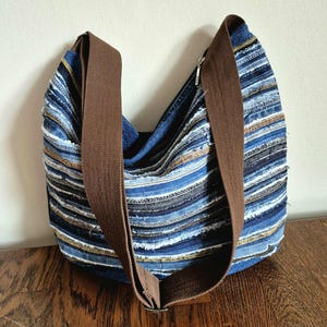 May include: A blue denim shoulder bag with a brown strap. The bag is made from strips of denim in various shades of blue, with accents of white and gold. The strap is a solid brown color.