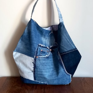 Peut inclure: Un grand sac fourre-tout en denim fabriqué à partir de jeans recyclés. Le sac présente un design patchwork avec différentes nuances de denim bleu, dont une poche avant. Le sac a une seule bandoulière.