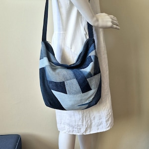 Könnte beinhalten: Eine Hobo-Umhängetasche aus einem Patchwork verschiedener Jeansstoffe in den Farben Dunkelindigo, Mittelblau und Hellblau. Die Tasche verfügt über einen breiten, integrierten Schulterriemen und eine geschwungene, lockere Silhouette.