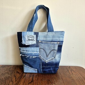 May include: A tote bag constructed from patchwork denim in various shades of blue. The bag features two denim handles, a visible back pocket, and a white rectangular Levi Strauss & Co. label with black text reading For over 140 years.