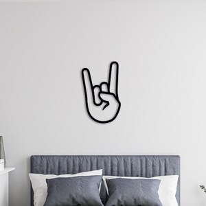 May include: Black metal wall decor in the shape of a hand making the rock and roll hand gesture. The decor is mounted on a white wall above a gray bed with white pillows. The bed is in a bedroom with a desk and chair, a lamp, and a plant.