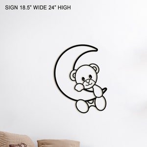 May include: A white metal wall decor featuring a teddy bear sitting on a crescent moon. The decor is 18.5 inches wide and 24 inches high. The decor is hanging on a white wall in a child's bedroom. The bedroom also features a white bed with brown bedding, a white nightstand with a woven basket, a white desk with a white chair, and a white shelf with a wooden abacus, a gray woven basket, and a framed print of the solar system.