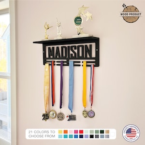 May include: A black metal medal display with the name "MADISON" cut out. The display holds several medals with colorful ribbons. Above the name are trophies. The text "21 COLORS TO CHOOSE FROM" is at the bottom.